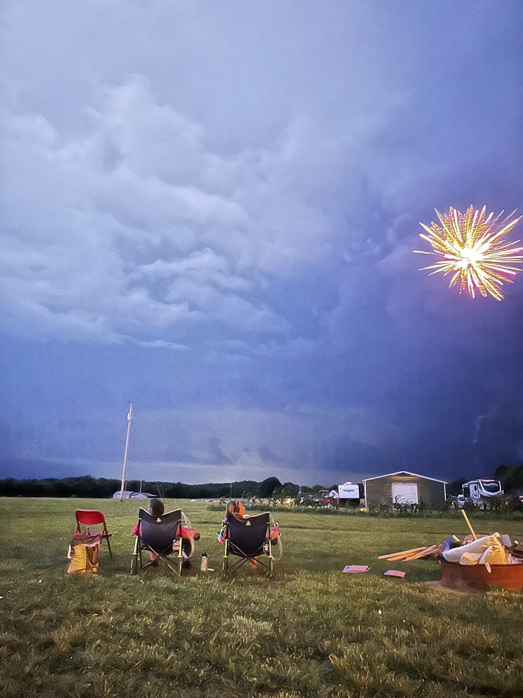 A single gold firework bursting over the field beneath heavy storm clouds, two people watching from camp chairs